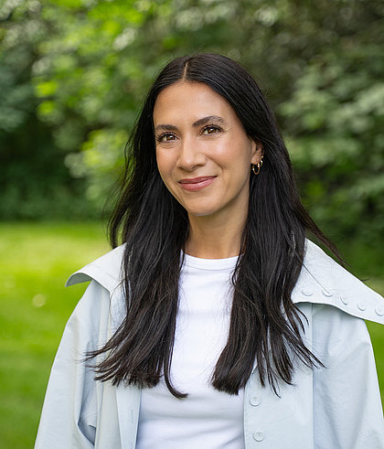Lächelnde Frau mit langen dunklen Haaren, hellblauer Jacke und weißem Shirt vor grünem Parkhintergrund.