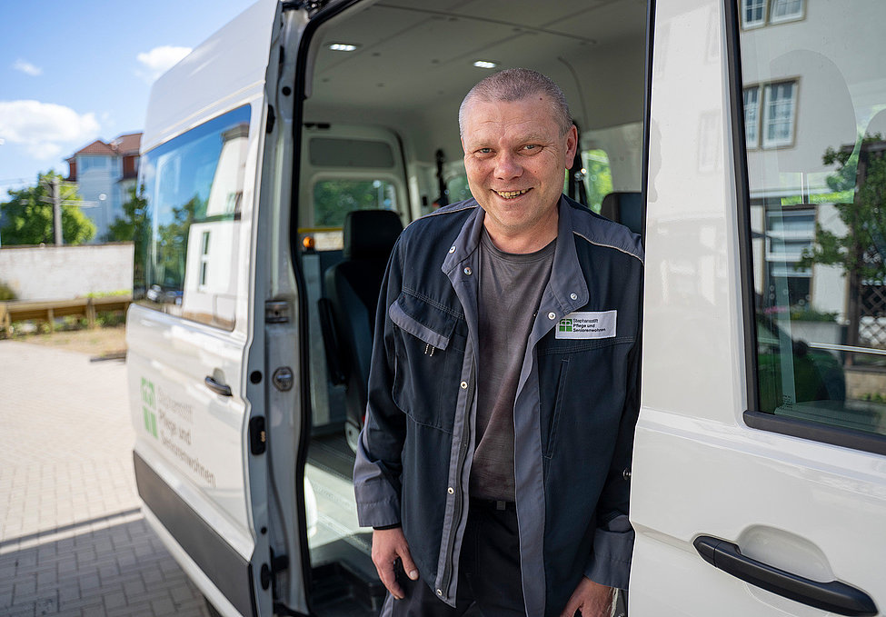 Ein Handwerker steht lächelnd in der offenen Tür eines Transporters, trägt Arbeitskleidung mit Namensschild und blickt direkt in die Kamera.
