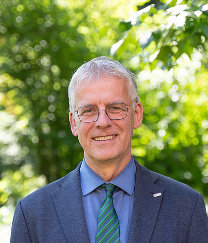Lächelnder Mann mit Brille, kurzen grauen Haaren und dunkelgrauem Anzug.