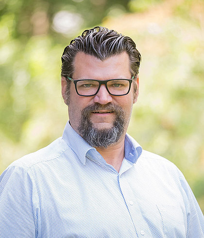 Mann mit Brille und Vollbart in hellblauem Hemd vor unscharfem grünen Hintergrund